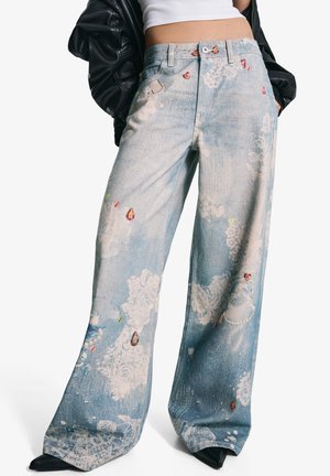 Wide-leg light blue jeans with white lace patterns and multicolored gem-like embellishments, worn with a cropped white top and black jacket.