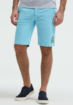 Mann trägt hellblaue Shorts bis zum Knie mit einem kleinen Aufnäher am linken Bein, kombiniert mit marineblauen Turnschuhen und einem gestreiften Hemd.