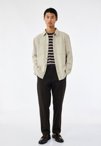 Joven con las manos en los bolsillos, vistiendo una chaqueta de camisa beige abotonada sobre una camisa a rayas en blanco y negro, con pantalones negros y zapatos oscuros.