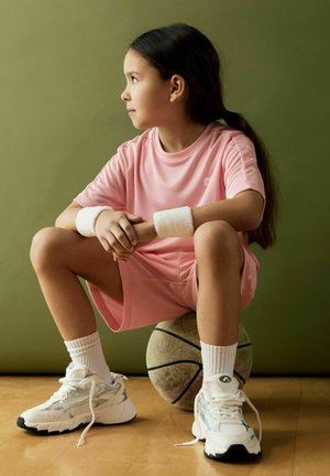 Ung jente i rosa treningsklær og hvite joggesko som sitter på en basketball og ser til siden mot en ensfarget grønn bakgrunn.