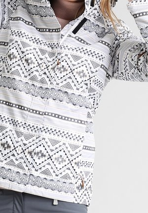 Vrouw draagt een wit jack met zwart- en grijsgekleurde geometrische patronen en een ritssluiting aan de voorkant, gecombineerd met grijze broek.