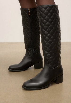 Bottes noires montantes jusqu’au genou avec dessus en cuir matelassé et dessous lisse, dotées d’une fermeture éclair latérale et de petits talons blocs, portées sur un sol beige.