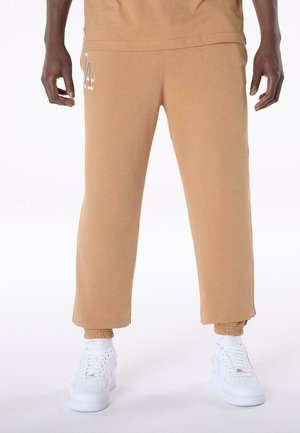 Person, die beige Sweatpants mit elastischen Bündchen und weißen Sneakern trägt, steht vor einem schlichten weißen Hintergrund.