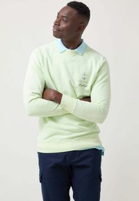 Sudadera verde claro con cuello redondo, mangas largas y puños de canalé. Presenta un logo de ancla y un cuello azul claro en varias capas. Pantalones azul marino.