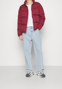 Bordeaux pufferjack met een ritssluiting, gedragen over een wit T-shirt, gecombineerd met lichtblauwe wijde jeans en marineblauwe sneakers met beige accenten.