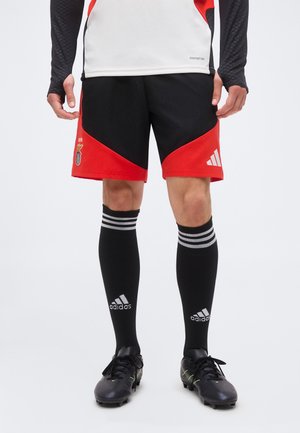 Homme portant un short de sport noir et rouge, des chaussettes montantes noires Adidas et des crampons noirs, debout avec les mains détendues le long du corps.