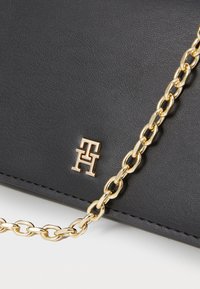 Sac en cuir noir avec une bandoulière en chaîne dorée et un accent logo doré. La texture est lisse avec un grain subtil.