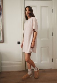 Roze oversized jurk met korte mouwen, gestructureerde stof en afgeronde zoom. Gecombineerd met bruine klompen en crèmekleurige sokken, staand op een houten vloer.