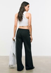 Zwarte wijde sweatpants met een trekkoord in de taille, gedragen met een witte cropped tanktop. Model houdt een gestreept shirt vast. Soepele textuur.