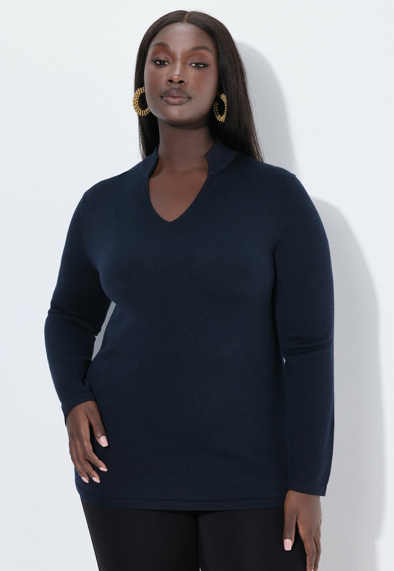 Navyblauw langemouwen gebreide top met een V-hals en geribbelde manchetten. Gladde textuur, rechte snit, gedragen over zwarte legging.
