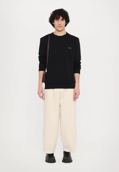 Sort sweatshirt med rund halsudskæring, prydet med farvede striber på brystet. Sammen med løstsiddende cremede bukser og sorte sko. Brun taske slidt over skulderen.