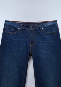 Jeans in denim blu scuro con tasche frontali, passanti per cintura e chiusura con bottone, mostrati dalla vita a metà coscia su sfondo chiaro.
