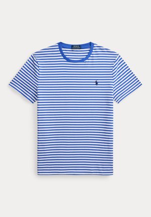 Jeune homme debout, les mains dans les poches, portant un t-shirt rayé bleu et blanc, un short bleu marine, des baskets blanches et une montre-bracelet.