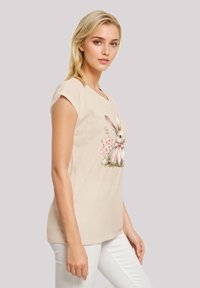 Beige cotton t-shirt with short sleeves featuring a rabbit illustration surrounded by pink flowers on the front. Smooth texture, casual fit.