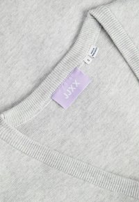 Lichtgrijze gebreide trui met een halslijn, voorzien van een lila JUX-label en kleine witte labels met maat S en het label 'gemaakt in Bangladesh'.