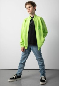 Fluorescency zielona kurtka zapinana na zamek z bocznymi kieszeniami, noszona na czarnej koszulce z grafiką, w połączeniu z jasnoniebieskimi dżinsami i czarnymi trampkami.