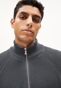 Grauer Strickpullover mit hohem Kragen, vollem Reißverschluss und geripptem Textur. Auffällige Details an den Schultern und das Stickmuster heben das Design hervor.