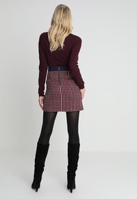 Viininpunainen pitkähihaisen yläosa yhdistettynä kuvioituun minihameeseen, jossa on punaisen ja laivastonsinisen sävyjä. Mustat reisiin ulottuvat saappaat täydentävät asun.