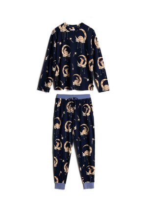 Femme portant un pyjama bleu marine assorti avec des motifs beiges de lune, étoile et lapin, ajustant le bas du haut du pyjama.
