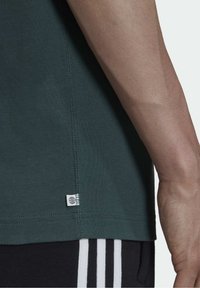 Nahaufnahme eines Arms einer Person neben dem Saum eines dunkelgrünen Shirts mit einem kleinen weißen Etikett und schwarzen Hosen, die drei weiße vertikale Streifen aufweisen.