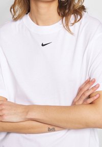 Camiseta de algodón blanca con cuello redondo y mangas cortas, que presenta un pequeño logo negro de Nike centrado en el pecho. Textura suave.