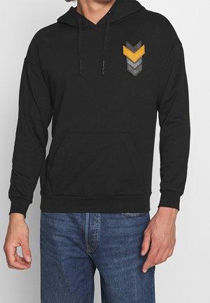 Sudadera con capucha negra con un parche de flechas en naranja, gris y negro. Cuenta con un bolsillo frontal y cordones en la capucha. Tejido suave.