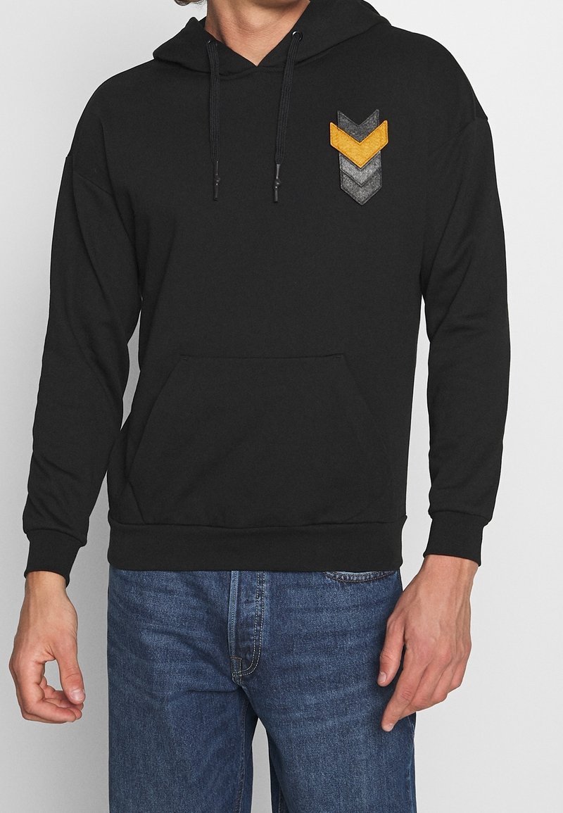 Felpa nera con cappuccio e un patch a chevron arancione, grigio e nero. Presenta una tasca frontale e lacci sul cappuccio. Tessuto morbido.
