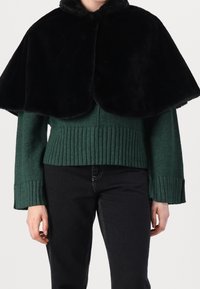 Grön stickad tröja med ribbad nederkant, med ett svart capeöverdrag i fejkpäls. Plagget har vida ärmar och rund halsringning.