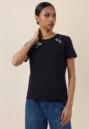 Mujer con el cabello peinado hacia atrás, que lleva una camiseta negra con adornos de pedrería cerca de los hombros y vaqueros de mezclilla azul.