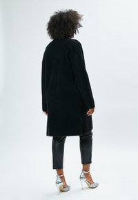 Cappotto lungo nero e soffice con spalle scese, indossato sopra pantaloni neri lucidi e scarpe con tacco alto argento. Design minimalista senza chiusure visibili.