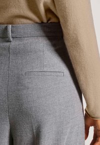 Graue maßgeschneiderte Hose aus strukturiertem Stoff, mit einer einzelnen Gesäßtasche und einem flachen Bund. Kombiniert mit einem beigen Pullover.