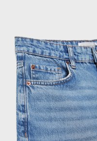 Vaqueros de mezclilla azul claro con una textura ligeramente descolorida, que presentan un bolsillo delantero, remaches de cobre y costuras naranjas a lo largo de las costuras.