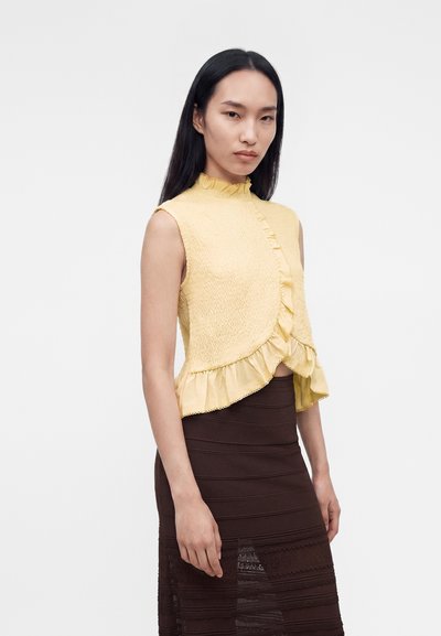Femme aux longs cheveux noirs portant un haut jaune sans manches à volants et une jupe texturée marron foncé taille haute, debout devant un fond uni.