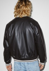 Veste aviateur en cuir noir avec poignets et taille côtelés rayés blanc et noir. Texture lisse, épaules arrondies et détails minimaux.