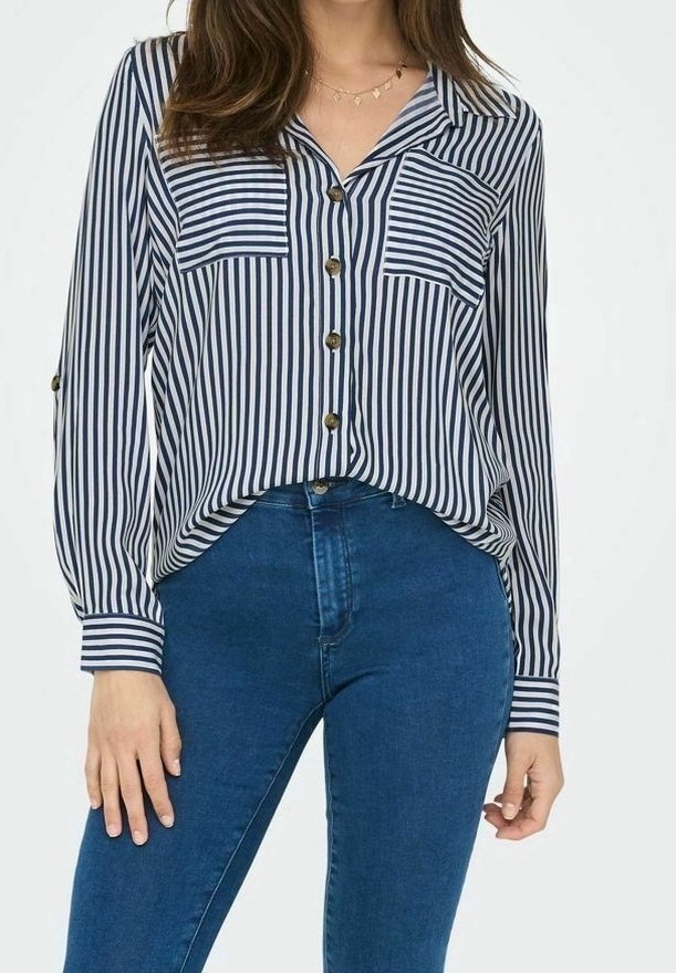 Camisa de botones con rayas verticales en azul marino y blanco, dos bolsillos en el pecho, mangas largas y un corte relajado, combinada con jeans azules.