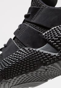 Black athletic shoe featuring a textured rubber sole, mesh upper, and suede accents, with elastic straps for secure fit and modern design elements.