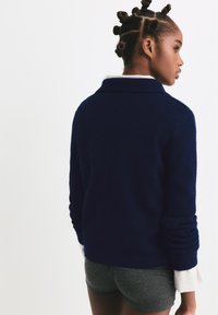 Cardigan blu navy con collo a scialle, polsini a costine e una textura morbida. Indossato sopra una camicia bianca a righe blu e pantaloni corti grigi.