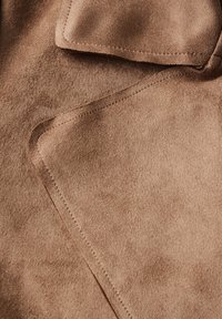 Primer plano de tela de ante marrón con costuras visibles y bordes doblados superpuestos, destacando la textura y los detalles de las costuras.