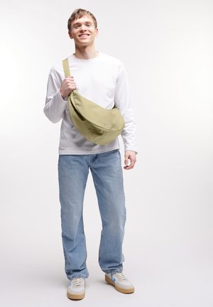Giovane uomo sorridente, vestito con una camicia bianca a maniche lunghe, jeans azzurri chiari, sneaker bianche e che tiene una grande borsa a tracolla verde oliva.