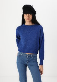 Suéter azul cortado com um tecido texturizado e felpudo, gola redonda e punhos canelados, combinado com jeans claros justos.