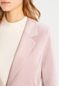 Kvinna i en ljusrosa blazer över en krämfärgad ribbad polotröja, med löst ljusbrunt hår synligt.