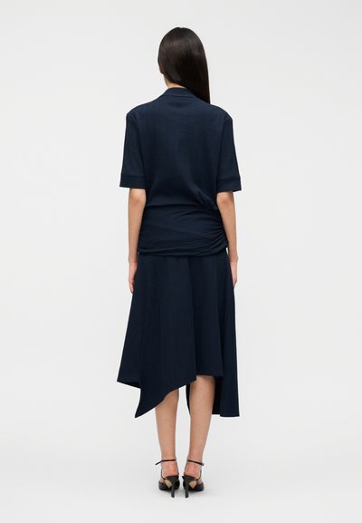 Φόρεμα σε ναυτικό μπλε χρώμα με άνετη εφαρμογή, κοντά μανίκια και ασύμμετρο τελείωμα. Διαθέτει λεπτομέρειες με πτυχώσεις στο πίσω μέρος, κατασκευασμένο από υφασμάτινη υφή.
