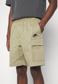 Shorts cargo kaki avec taille élastique, poches latérales et poche zippée. Présente un logo Nike noir sur la jambe. Matière en coton.