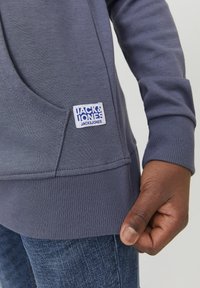 Sudadera con capucha gris de acabado texturizado, que cuenta con un bolsillo frontal, puños acanalados y una etiqueta de tela con el logo "JACK & JONES".