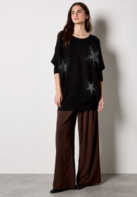 Czarny oversize sweter z srebrnymi zdobieniami w kształcie gwiazdek oraz kropkowymi wzorami, zestawiony z luźnymi brązowymi spodniami w szerokim kroju i czarnymi botkami za kostkę.