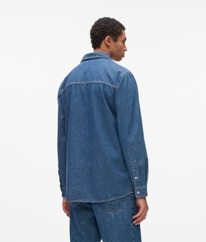 Bărbat purtând o jachetă din denim albastru și blugi, stând cu spatele către cameră, pe un fundal alb simplu.