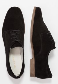 Scarpe oxford in suede nero con texture liscia, punta rotonda e lacci. Suola in gomma marrone chiaro con dettagli sottili sui lati.