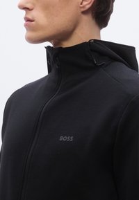 Sudadera negra con cremallera y cuello alto, tejido texturizado y un logo sutil impreso. Cuenta con cordones ajustables en la capucha para un mejor ajuste.