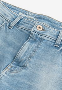 Svijetloplave traperice s kopčanjem na gumb i detaljima šivanja. Ima oznaku "DENIM GOODS CO" na pojasu.