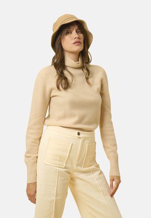 Beige, ribbet turtleneck sweater parret med lysegule, vide bukser med store påsyede lommer. Modellen bærer en matchende bucket hat.